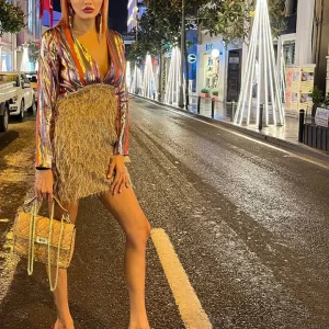 yakası açık parlak kumaş saçak detaylı etek ile birleşmiş tek parça mini karla elbise ile gece caddede poz veriyor